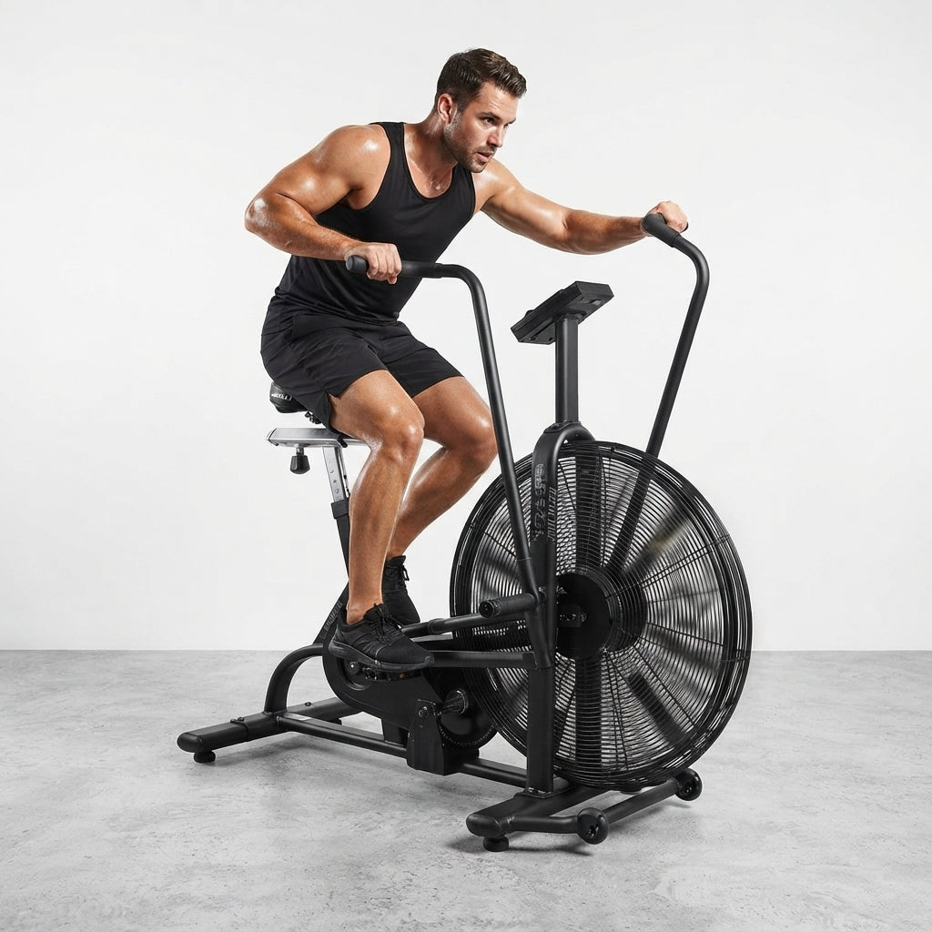 A man in a black tank top and shorts rides a black air resistance exercise bike with a large front fan wheel in a bright, minimalist studio setting.
